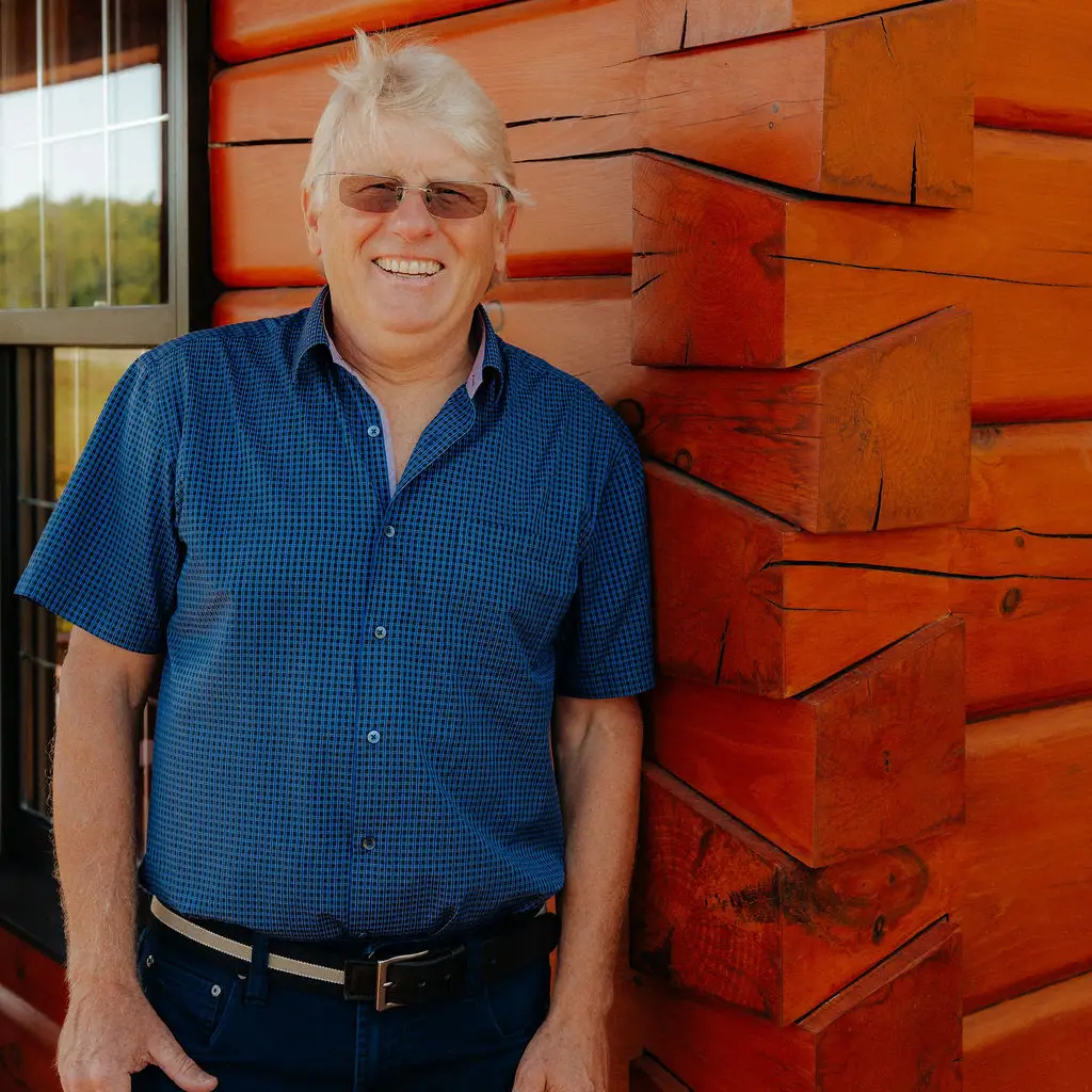 A man standing beside a wooden log cabin wall, smiling and wearing a blue checked shirt.